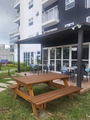 Uma área de estar coberta ao ar livre, com mesa e cadeiras de madeira, localizada ao lado de um edifício com janelas amplas e varandas.