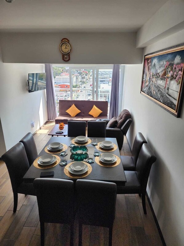 Uma área de refeições com uma mesa posta para seis pessoas, um sofá com almofadas, um relógio e um quadro na parede.