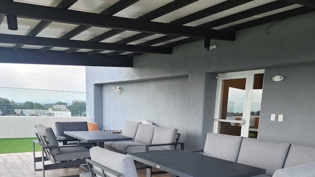 An outdoor patio with a long table and multiple chairs, a covered ceiling, and a view of the cityscape.