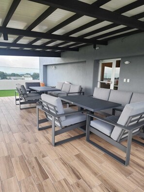 Um pátio externo com uma mesa comprida e várias cadeiras, teto coberto e vista para o horizonte urbano.