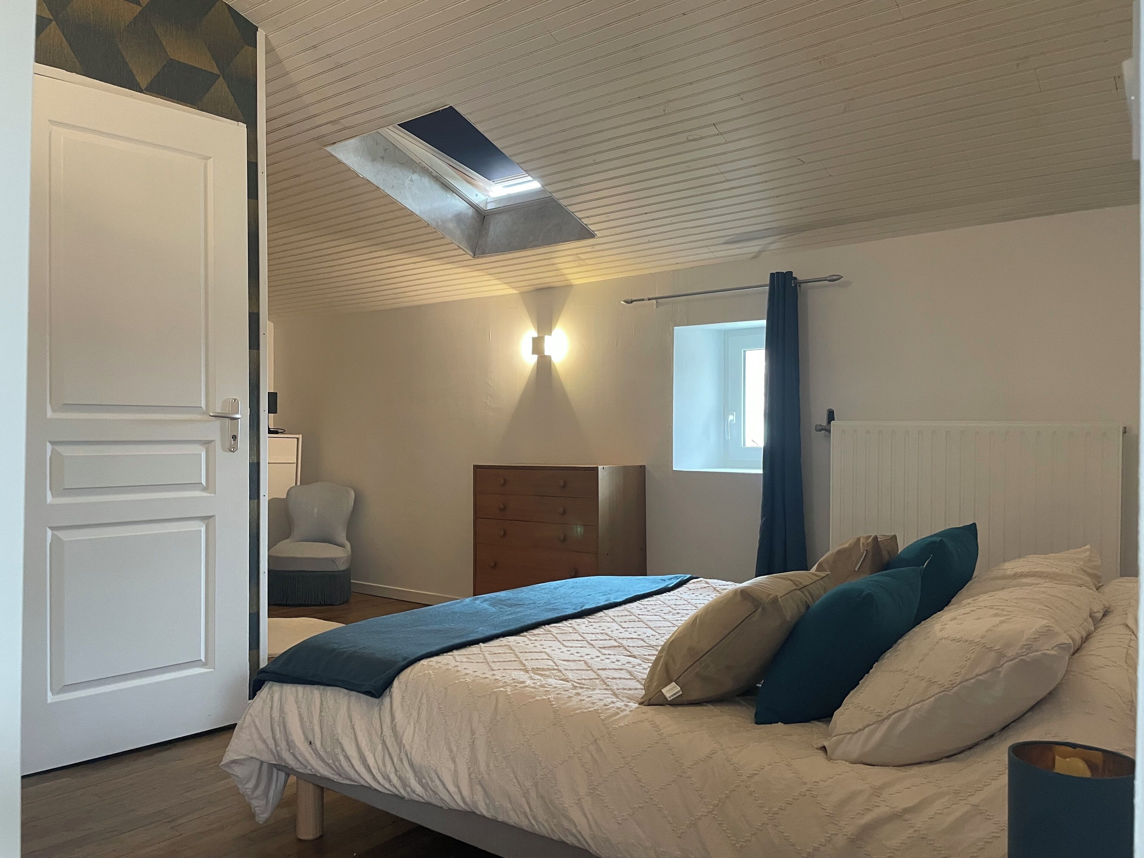 Een slaapkamer met een bed, een stoel, een dressoir, een raam met gordijnen en een ventilatie rooster.