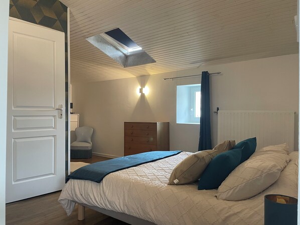 A bedroom with a bed, a chair, a dresser, a window with curtains, and a ceiling vent.