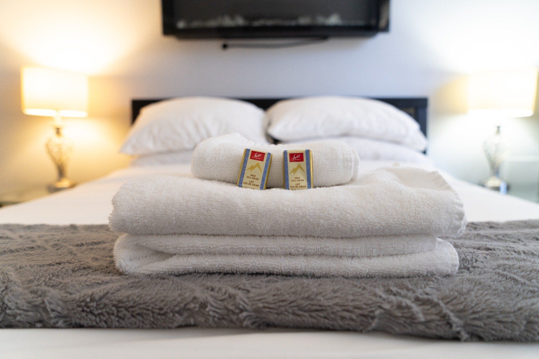 A neatly made bed with folded white towels and a gray blanket, a TV mounted on the wall, and two bedside lamps.