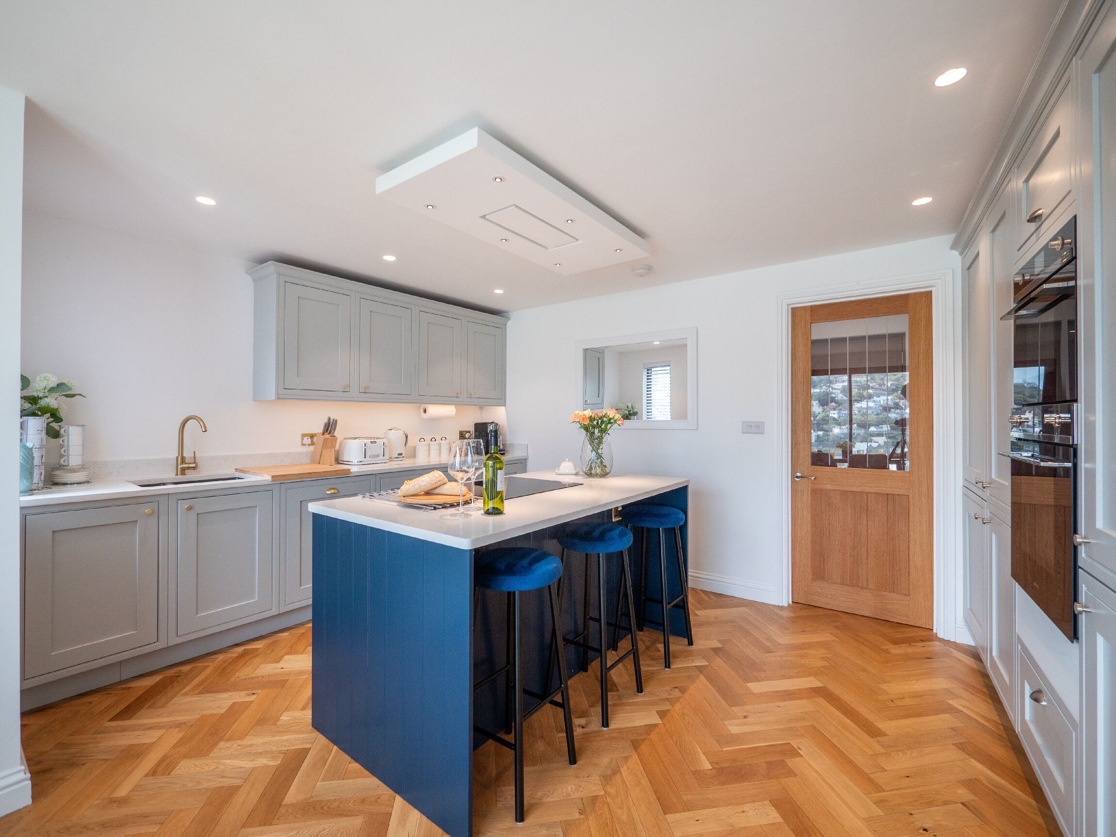 A modern kitchen with a central island, bar stools, and a view through a door.