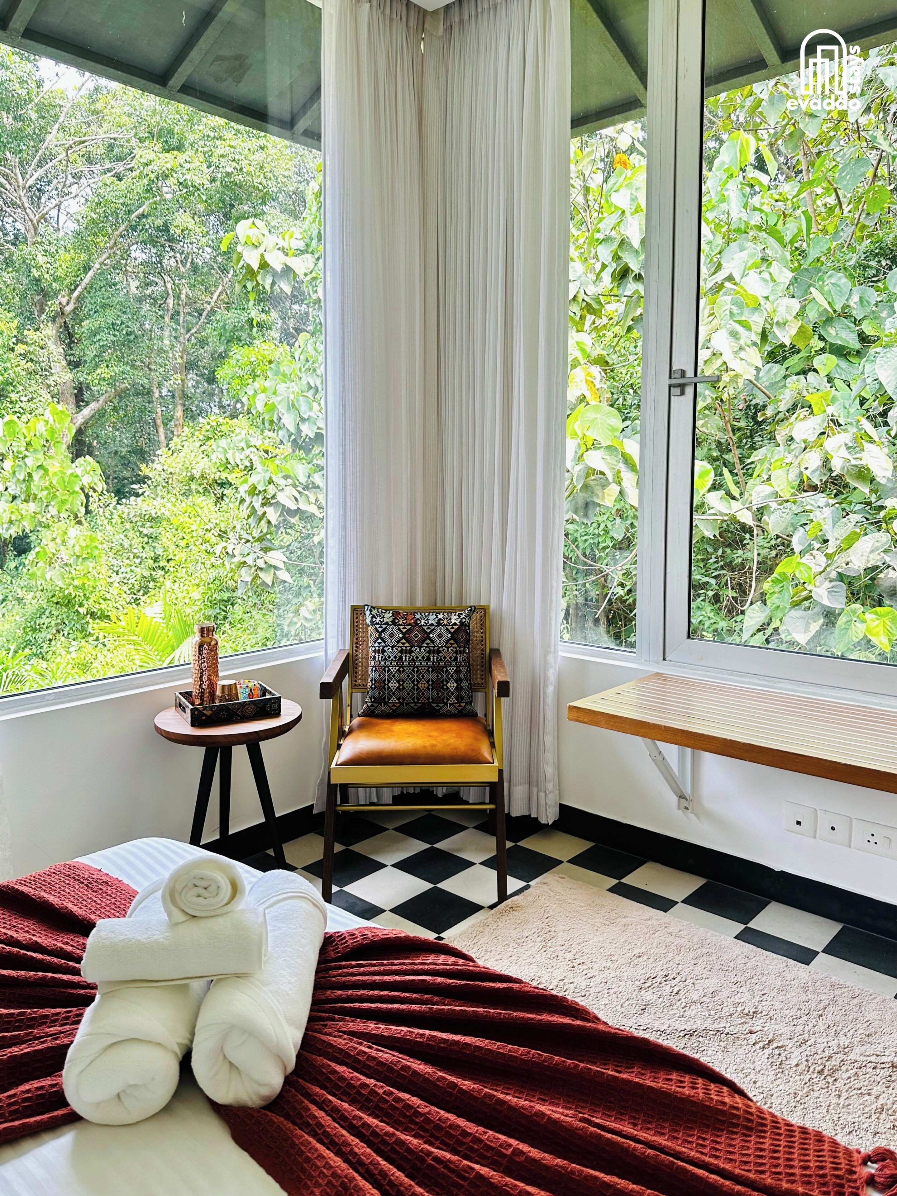 Una habitación con piso a cuadros, cama con manta roja, silla con cojín estampado, una mesita con un adorno y una ventana con vista a follaje.