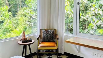 Una habitación con piso a cuadros, cama con manta roja, silla con cojín estampado, una mesita con un adorno y una ventana con vista a follaje.