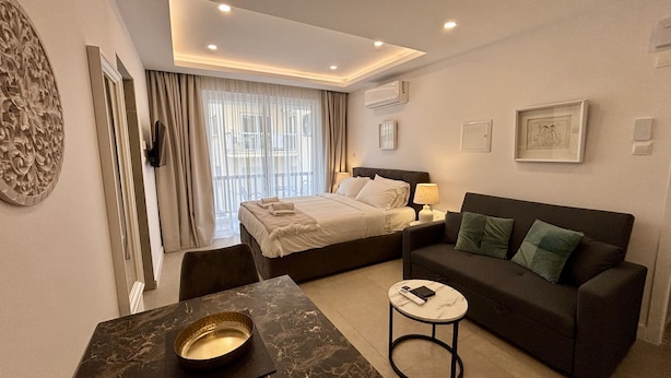 A modern hotel room with a bed, a sofa, a small table, and a decorative wall piece. - White Arches Studio Suite (Limassol)