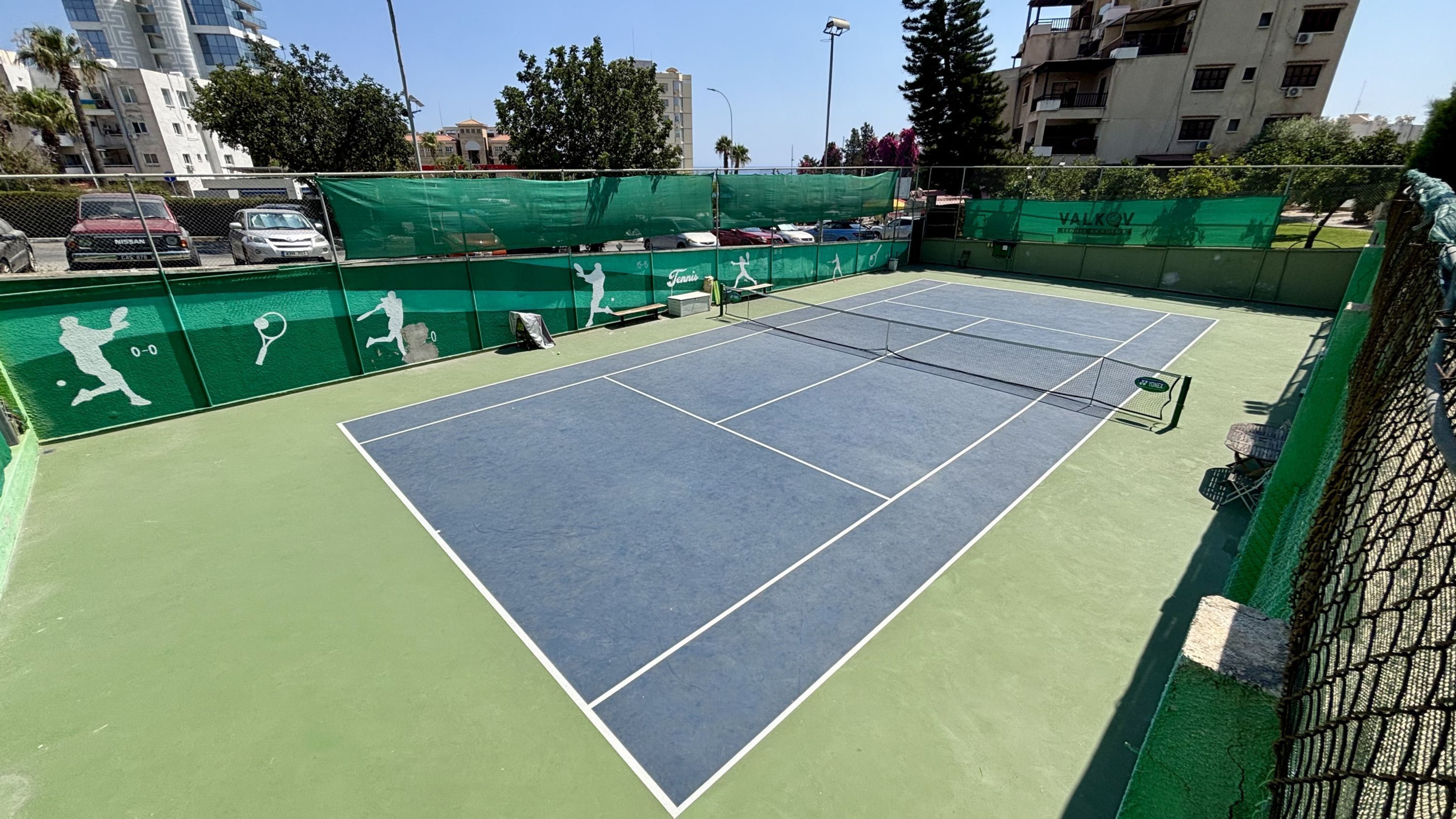 Un court de tennis aux surfaces vertes et bleues, entouré d’une clôture et d’arbres.