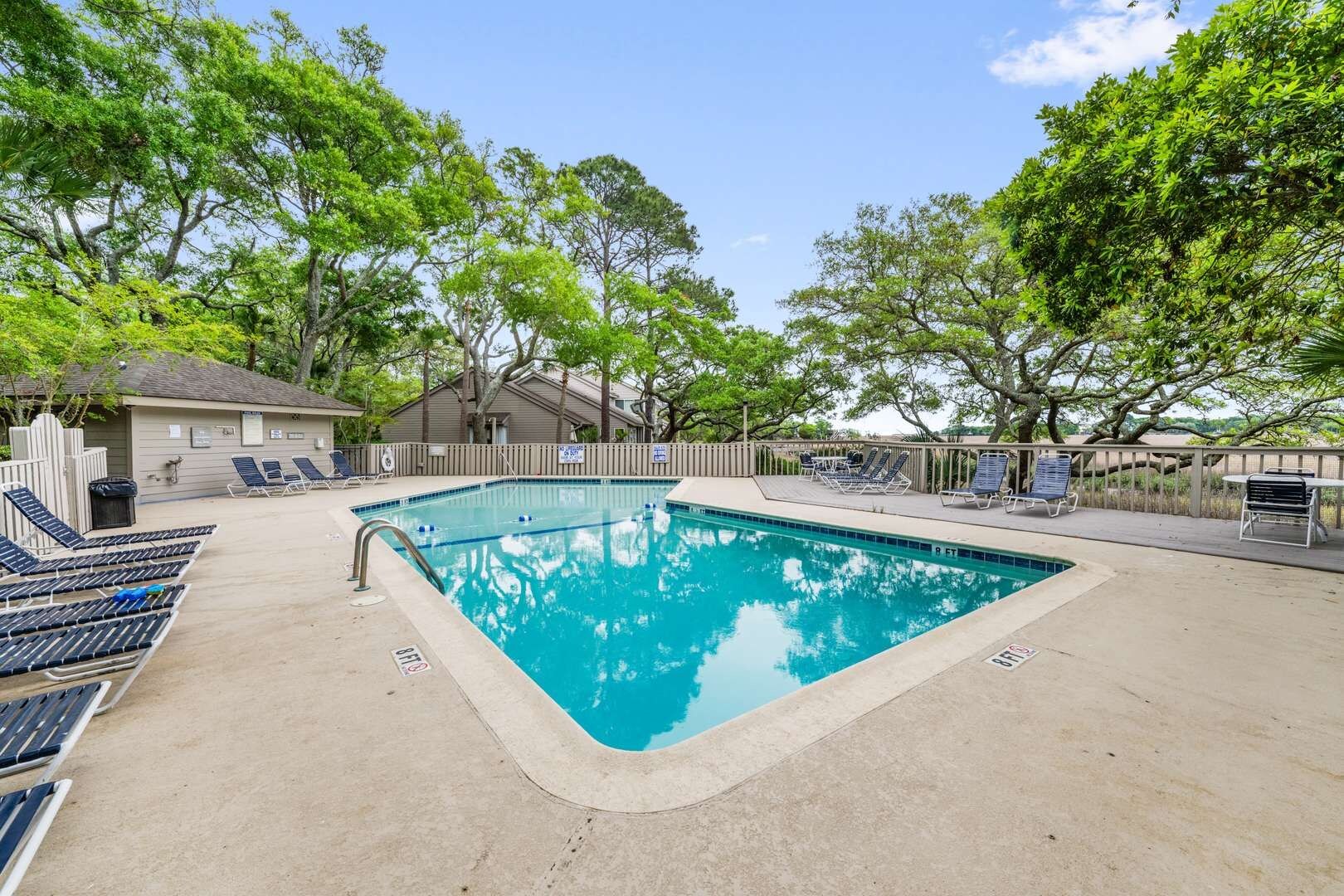 A swimming pool with lounge chairs and a covered area, surrounded by trees and a building.