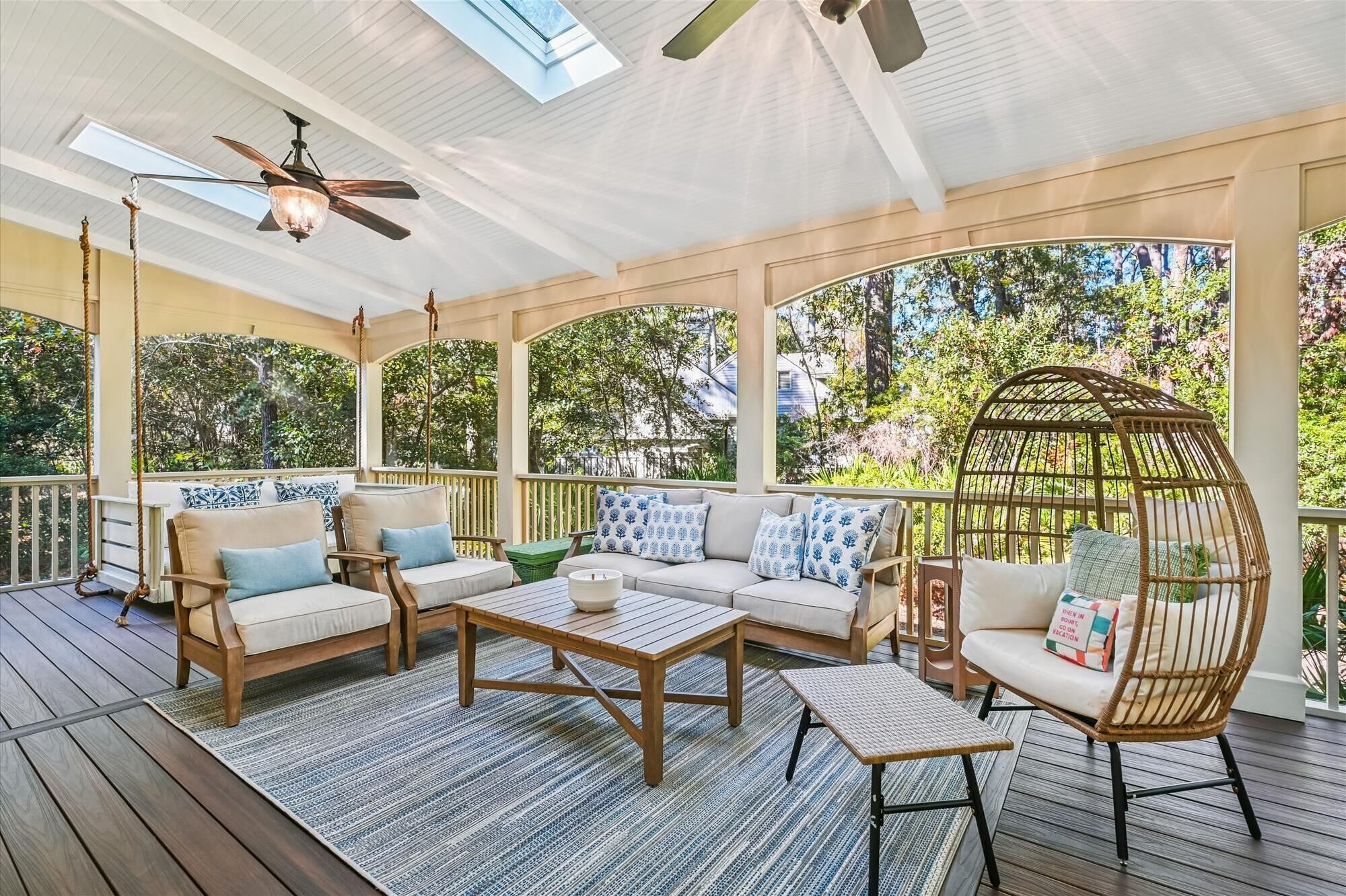 A covered outdoor seating area with a ceiling fan, wooden furniture, and a view of greenery.