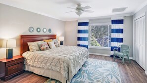 Ein Schlafzimmer mit Bett, Nachttisch, Lampe, Sessel, Fenster mit Vorhängen und Deckenventilator.