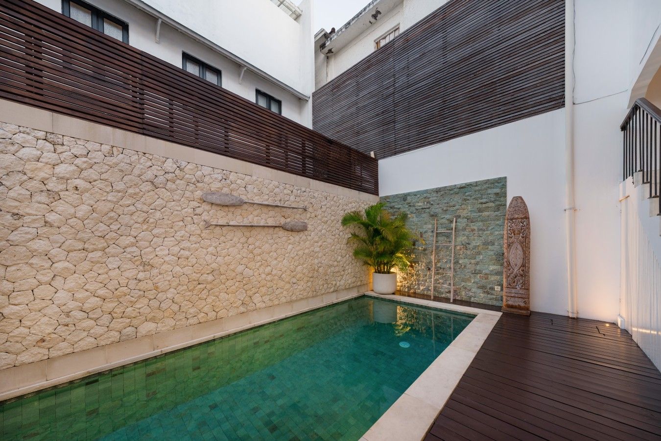 Une piscine avec des murs en pierre, une terrasse en bois et une plante en pot.