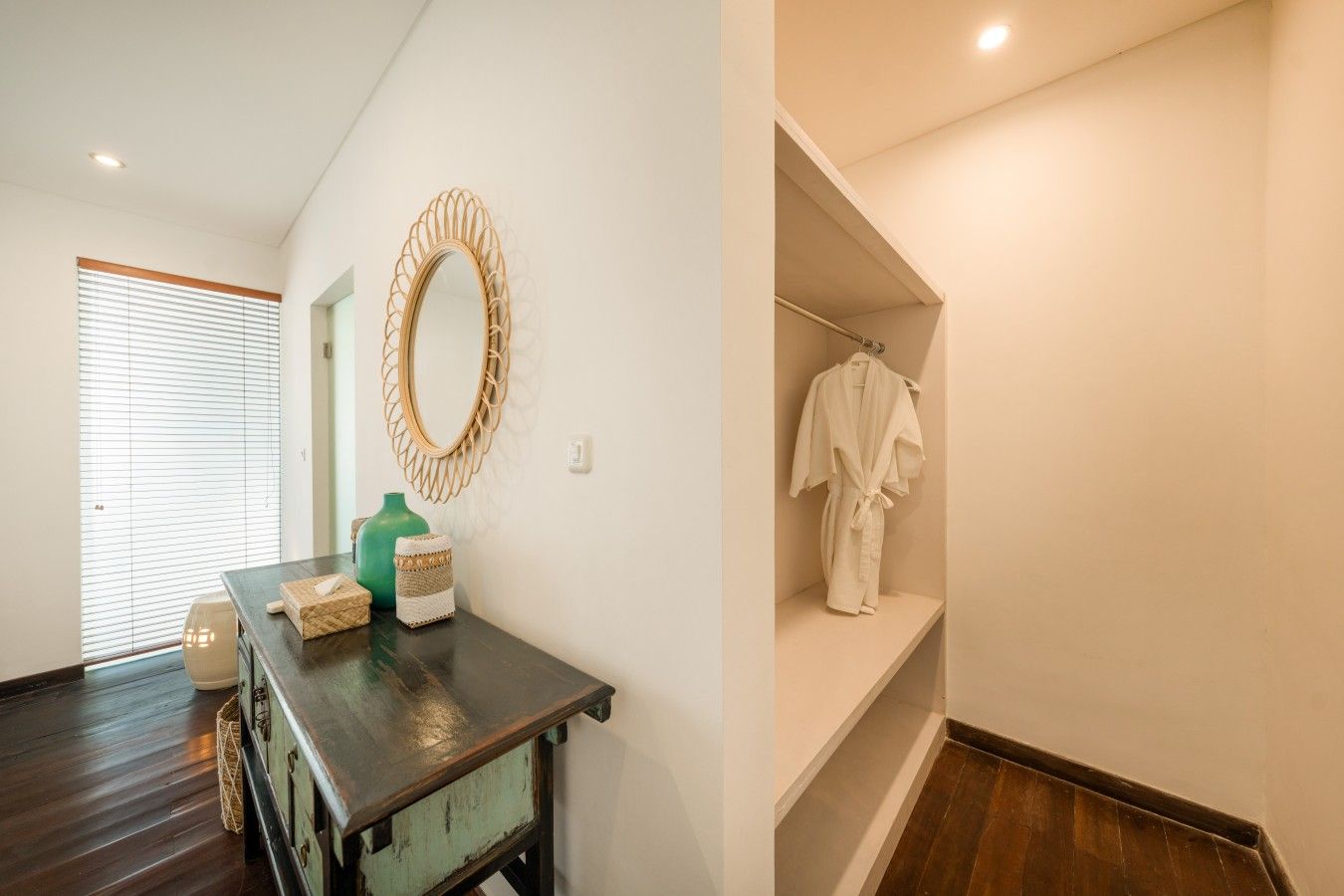 A room with a wooden floor, a dark wooden table with a mirror, a green vase, and a white robe on a shelf.