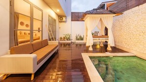 Un espacio exterior acogedor con terraza de madera, zona de estar con cojines, una pequeña piscina y un muro de piedra.