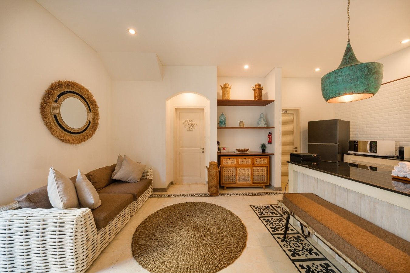 A living room with a sofa, a wicker chair, a round mirror, and a kitchen area with a refrigerator, microwave, and a hanging light fixture.
