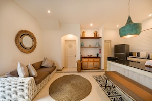 A living room with a sofa, a wicker chair, a round mirror, and a kitchen area with a refrigerator, microwave, and a hanging light fixture.