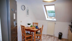 Una cocina compacta con una mesa de madera, sillas, un refrigerador, un reloj, una ventana y un radiador.