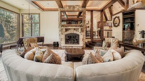 A spacious living room with a stone fireplace, large windows, and a high ceiling with exposed beams. - Immaculate Golf Course Home, Private Hot Tub, Gym, Firepits (Beaver Creek)