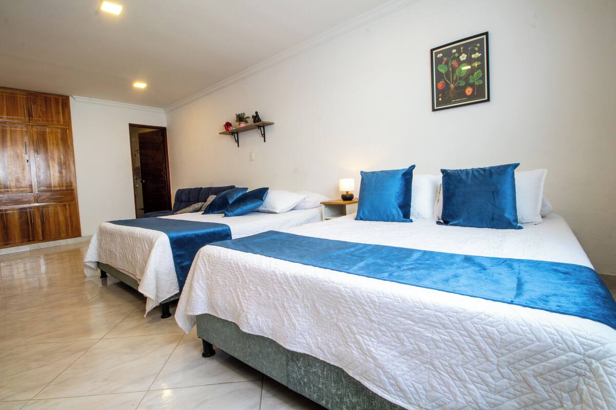 Una habitación de hotel con dos camas, un armario de madera, un estante en la pared con objetos decorativos e ilustración botánica enmarcada.