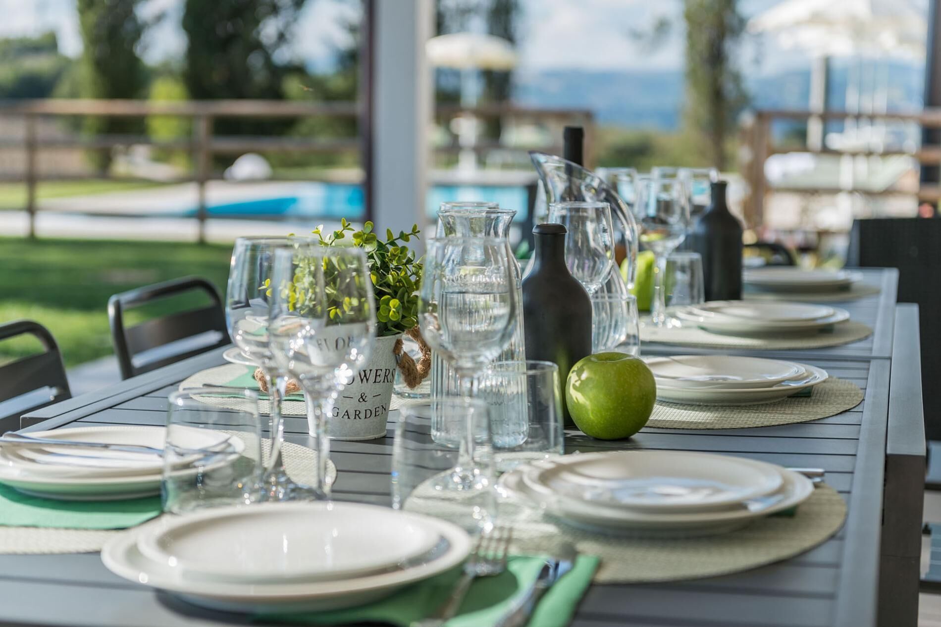 Tavola apparecchiata per un pasto, con piatti, bicchieri e una brocca, all'aperto con vista su una piscina e vegetazione.