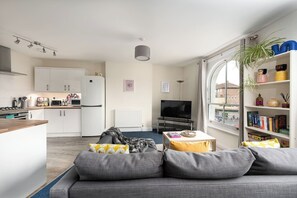 Een moderne woonkamer met een grijze bank, een klein tafeltje, een televisie en een boekenkast met diverse spullen.