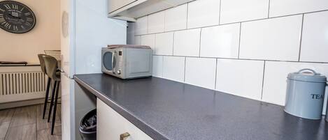 A kitchen with white cabinets, a microwave, a trash bin, and a clock on the wall.