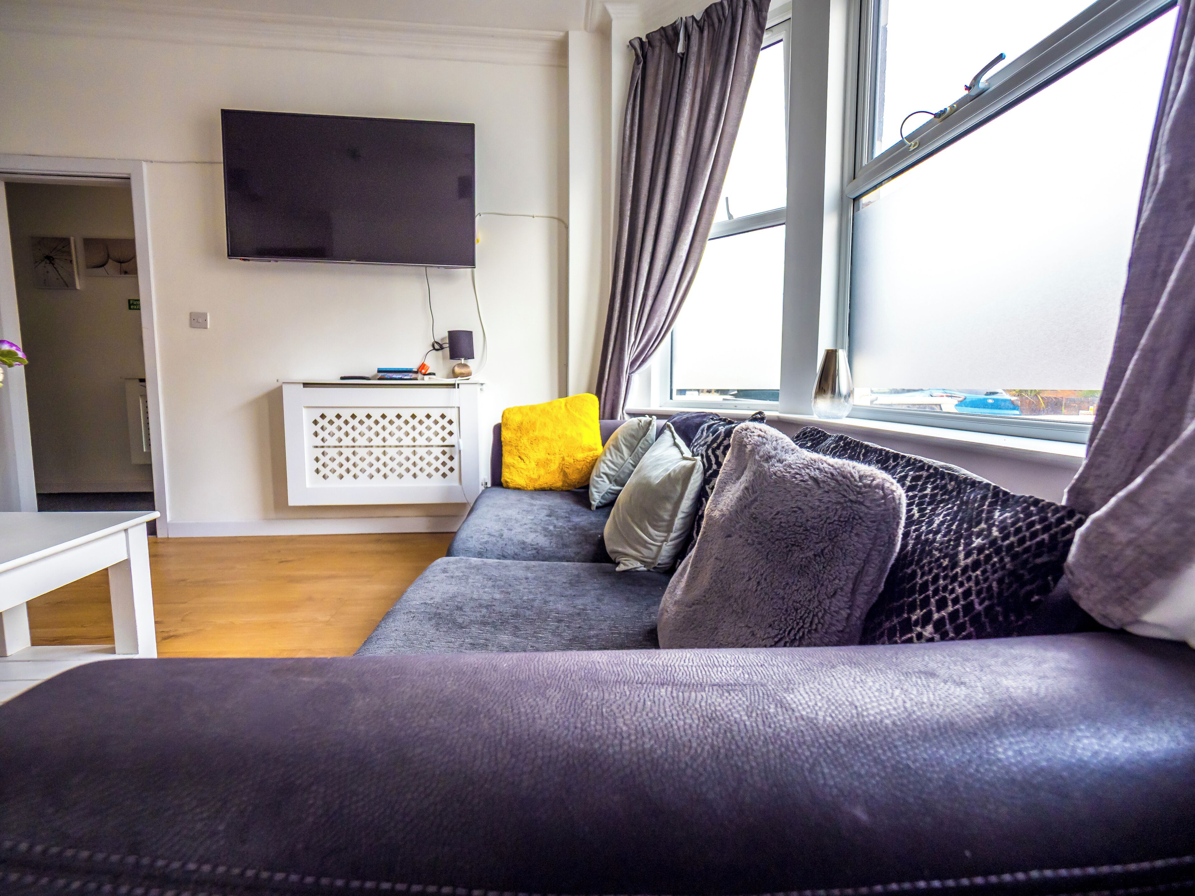 A living room with a grey sofa, a flat-screen TV mounted on the wall, and a window with curtains.