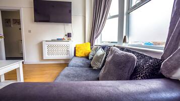 A living room with a grey sofa, a flat-screen TV mounted on the wall, and a window with curtains.