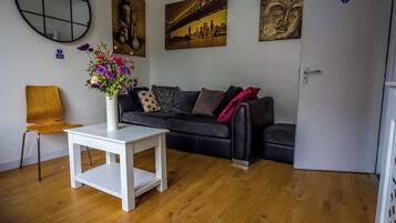 Sala de estar com mesa branca, sofá preto e um vaso de flores. Há quadros na parede e uma porta com uma placa.