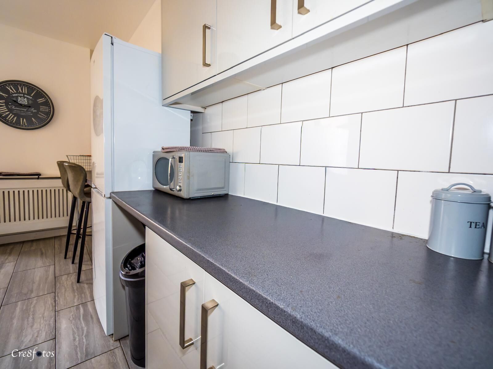 A kitchen with white cabinets, a microwave, a kettle, and a clock on the wall.