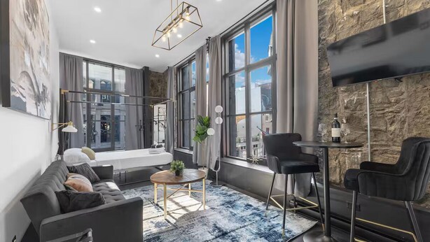 A modern living room with a sofa, a round coffee table, and a dining area with chairs. Large windows provide natural light and a view of the city. - AirStay-Saint Paul  (Montréal)