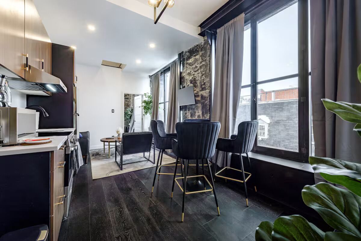 Eine moderne Küche mit dunklen Holzschränken, einer Edelstahl-Dunstabzugshaube und einem eingebauten Backofen. Vier schwarze Stühle mit goldenen Akzenten sind um einen kleinen Tisch angeordnet. Große Fenster mit Vorhängen sorgen für natürliches Licht und einen Blick auf die Stadt.