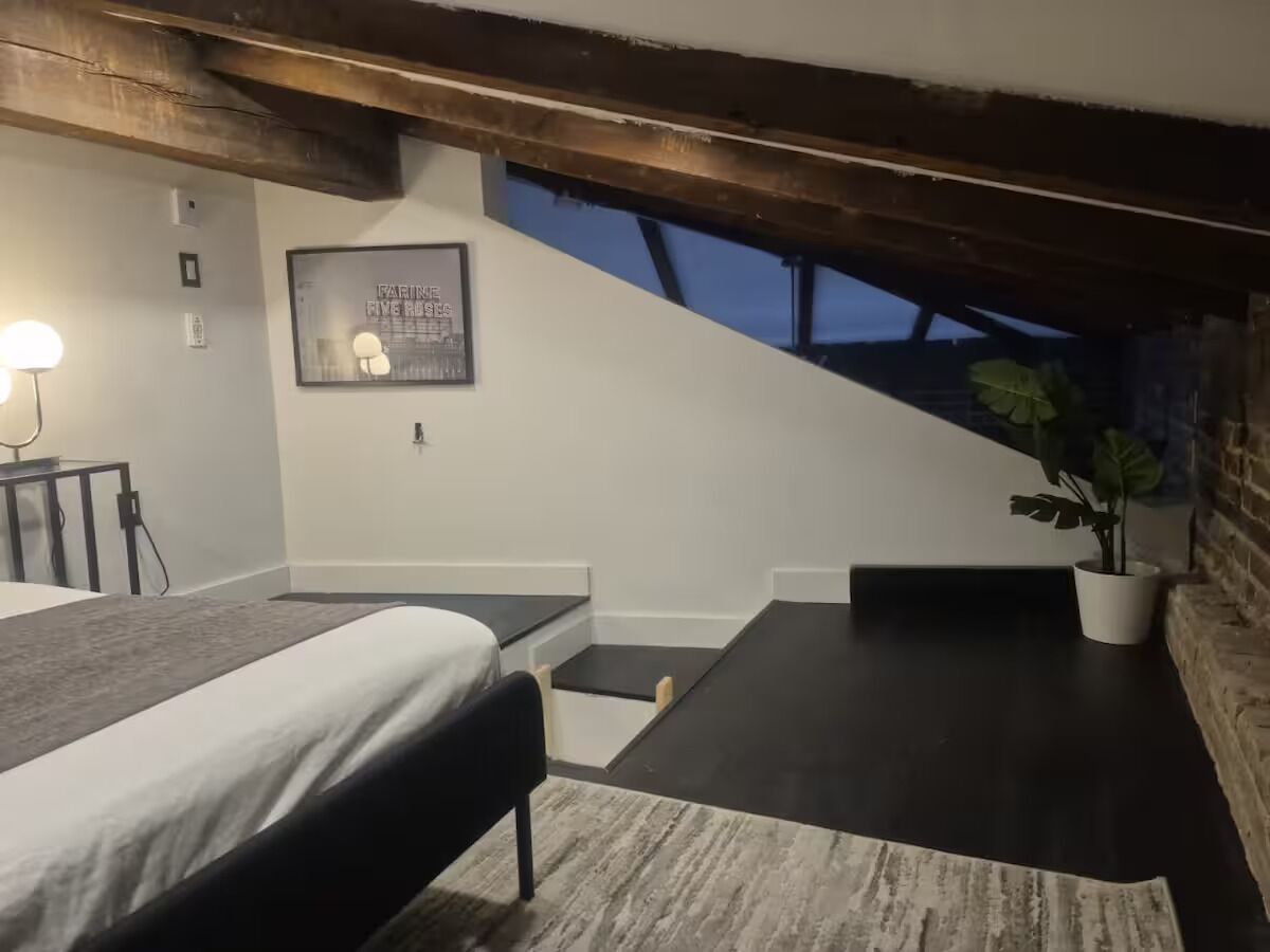 A bedroom with a bed, a desk, a chair, a small table, a potted plant, and a framed picture on the wall.