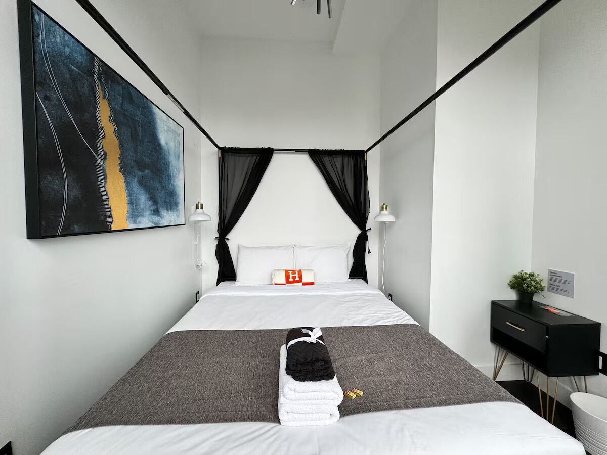 A modern hotel room with a large bed, black curtains, a bedside table with a lamp, and a painting on the wall.