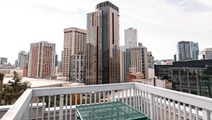 A rooftop terrace with green metal picnic tables and benches, overlooking a city skyline with various buildings.