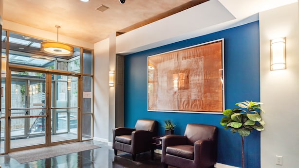 A lobby with a glass door entrance, two leather chairs, a potted plant, and a large abstract painting on the wall.