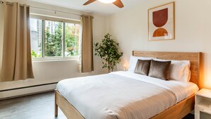 A bedroom with a bed, a ceiling fan, a window with curtains, a bedside table, and a wall-mounted artwork.