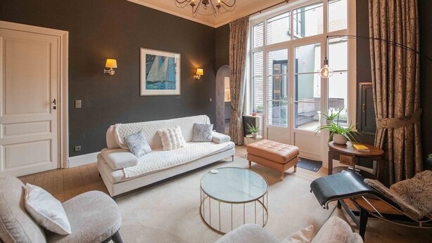 Een woonkamer met een witte bank, een glazen salontafel en een lange fauteuil. Er is een groot raam met gordijnen, een kroonluchter en wandlampen.