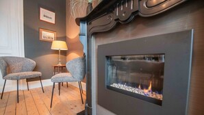 Een moderne woonkamer met een open haard, twee grijze fauteuils, een kleine tafel met een lamp en lijstwerken aan de muur.