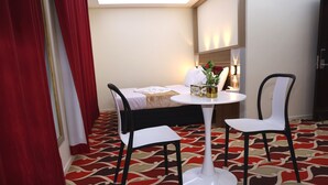 Habitación de hotel con una cama, una mesa redonda con dos sillas, una mesita con un jarrón y una planta, y una alfombra estampada.