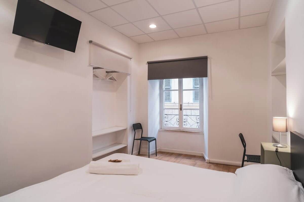 Ein Hotelzimmer mit einem Bett, einem an der Wand montierten Fernseher, einem Fenster mit Jalousien, einem Sessel und einem Nachttisch mit Lampe.