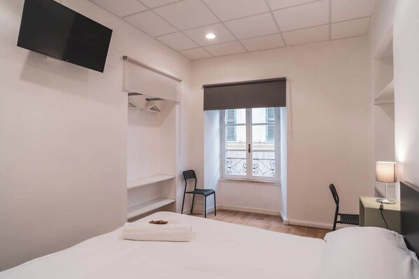 A hotel room with a bed, a TV mounted on the wall, a window with blinds, a chair, and a bedside table with a lamp.