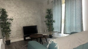 A living room with a sofa, a TV, a small table, two potted plants, and a window with curtains.