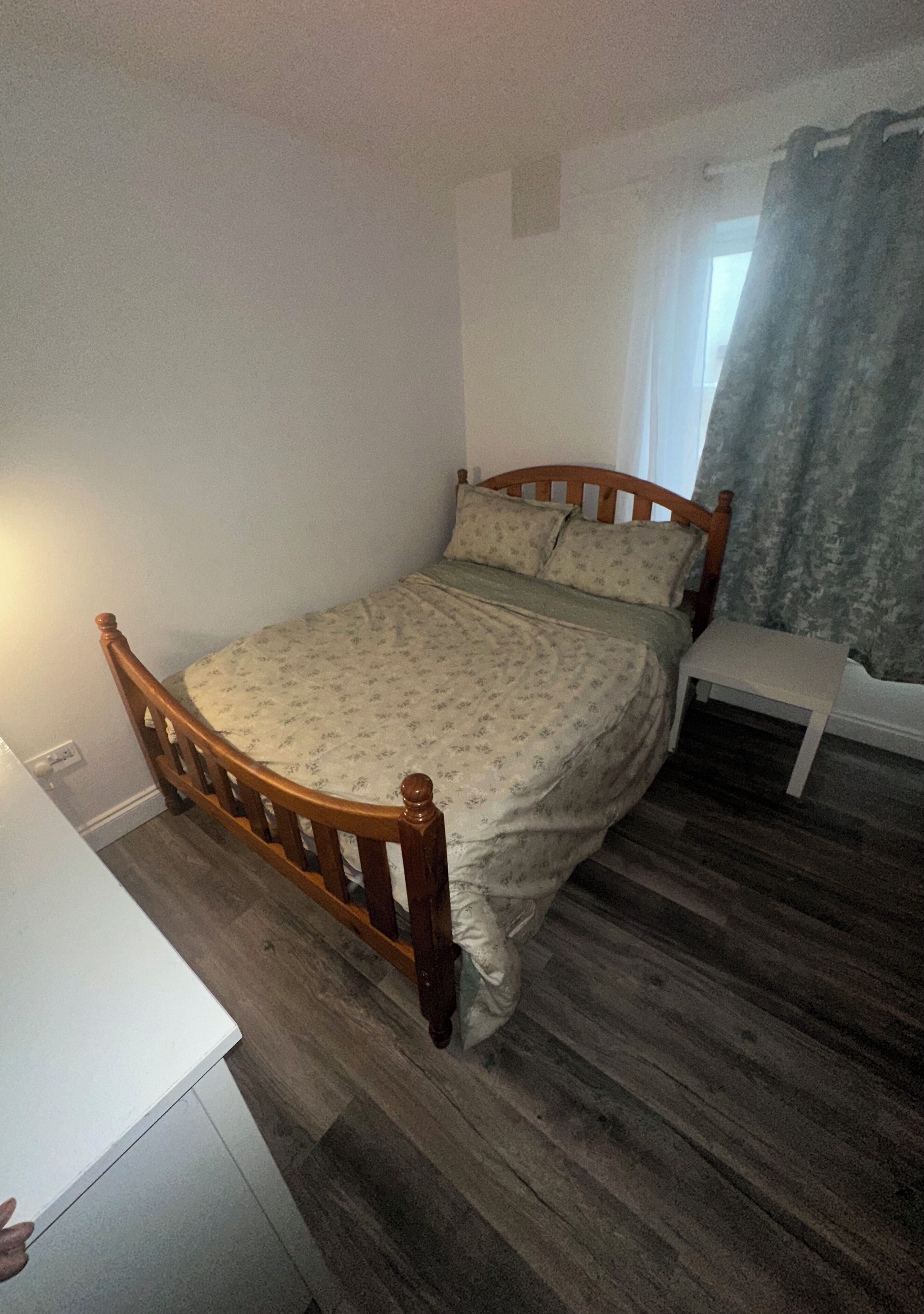 A single bed with a wooden frame and floral bedding in a room with wooden flooring and a window with curtains.