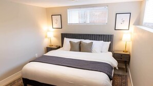 A neatly arranged bedroom with a large bed, two bedside tables with lamps, a window with blinds, and framed artwork on the walls.