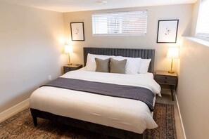 Quarto bem arrumado com uma cama grande, duas mesas de cabeceira com abajures, uma janela com persianas e quadros nas paredes.