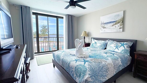 Ein Hotelzimmer mit einem großen Bett, einem Deckenventilator, einem Fernseher und Blick auf den Strand durch das Fenster.