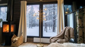 A cozy room with a wood-burning stove, a large window with a snowy forest view, a floor lamp, and a plush fur throw. - Riverside Village Ylläs (Kolari)