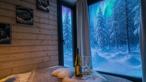 A cozy room with wooden walls, a window with a view of a snowy forest and aurora borealis, a bottle of wine, and two glasses on a furry rug. - Riverside Village Ylläs (Kolari)