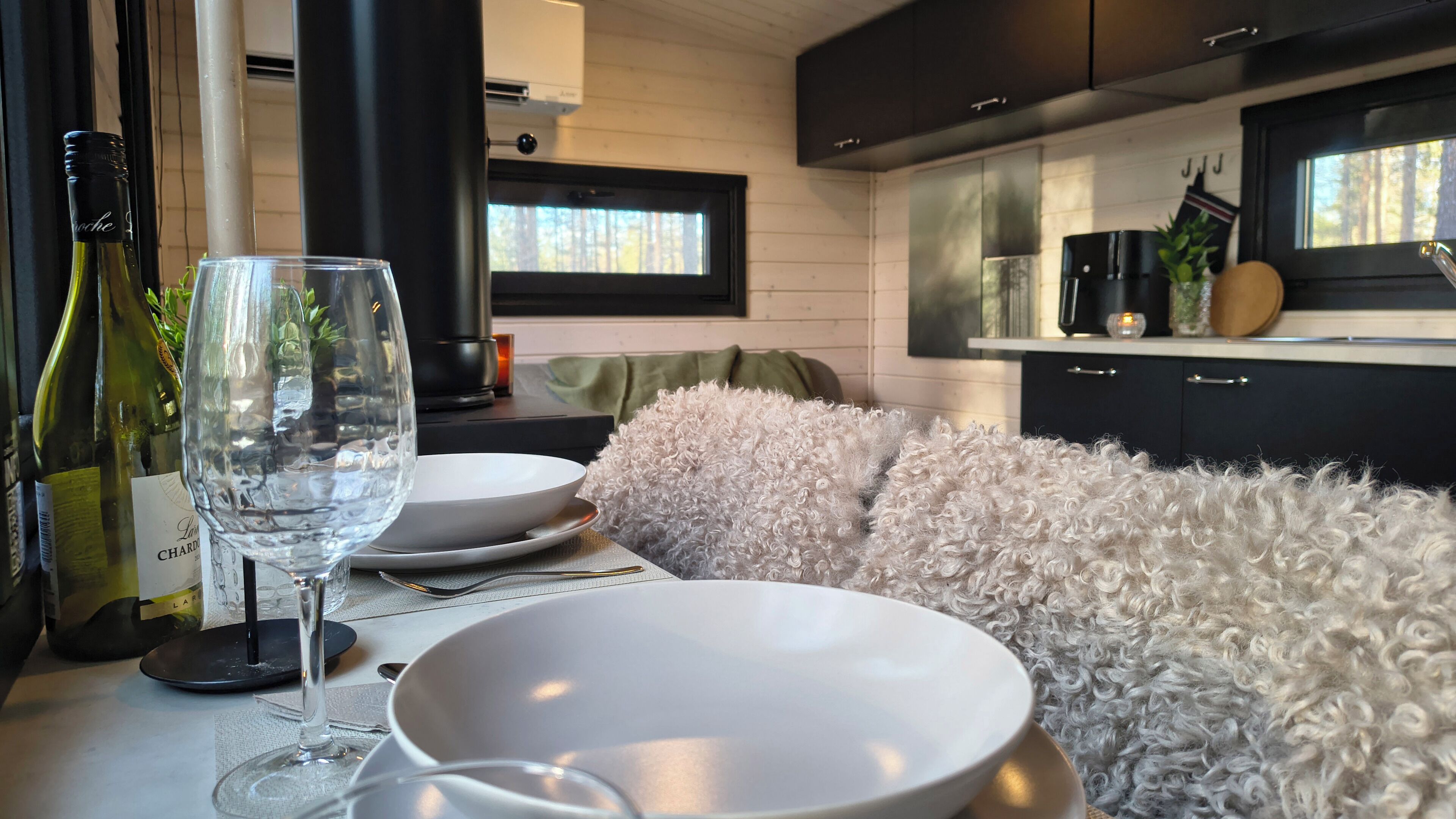 A modern kitchen with dark cabinets, a dining area with white plates, a fluffy white sofa, and a wine bottle on the table.